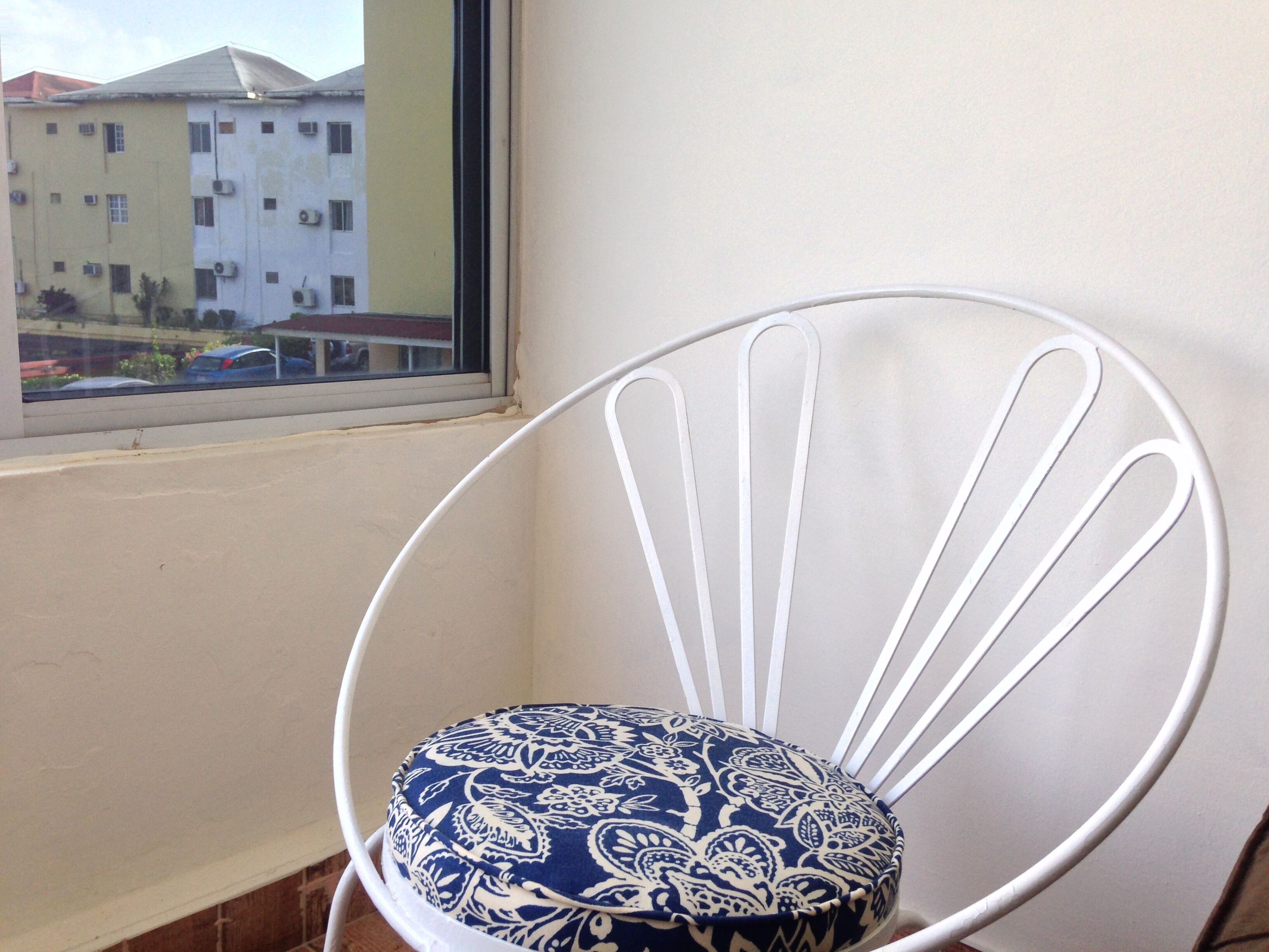 Uma cadeira de metal branca com almofada estampada em azul, posicionada perto de uma janela com vista para um edifício.