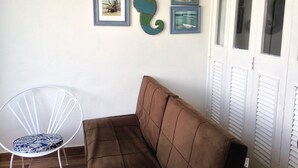 Een kamer met een bruine bank, een witte stoel met een gekleurd kussen, en drie ingelijste foto's aan de muur.