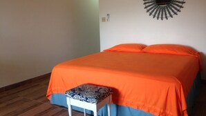 Een slaapkamer met een bed, een oranje sprei, een gekleurd kussen, een klein wit bankje en een spiegel aan de muur.