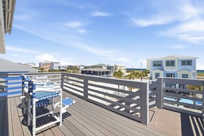 A wooden deck with a pool table and lounge chairs, overlooking a beach with buildings and a clear sky.