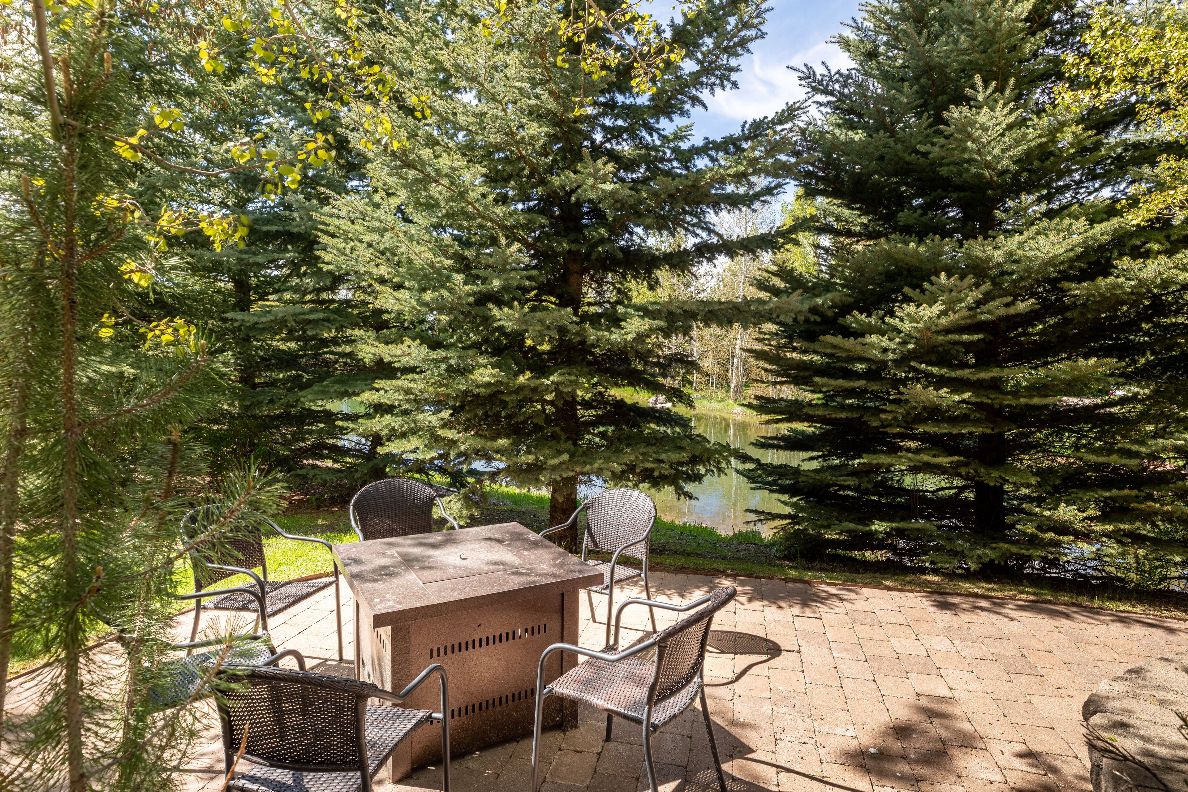 An outdoor patio area with a grill, chairs, and a view of a lake surrounded by evergreen trees.
