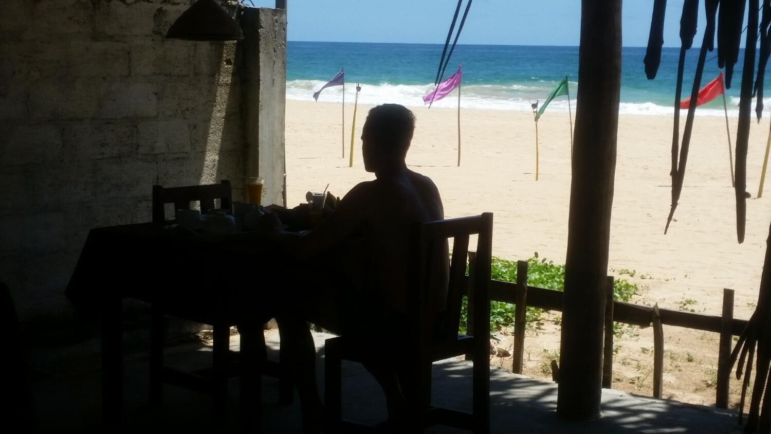 Vista desde la playa, desde un área sombreada con mesa y sillas, con vista al mar, banderas y una cerca de madera.
