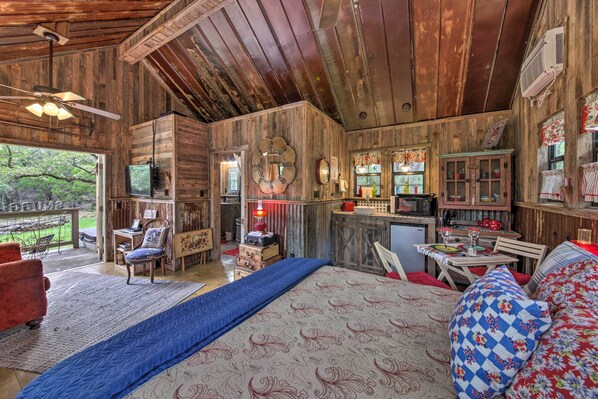 Interior rústico de una cabaña con cama, zona de estar, kitchenette y un amplio ventanal con vistas al exterior.