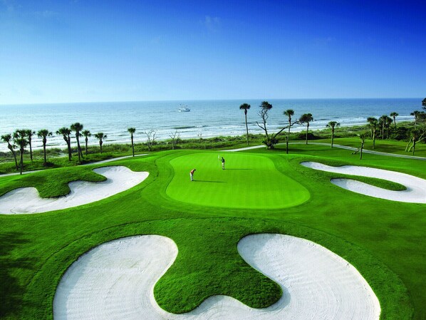 Un parcours de golf avec des bunkers de sable, une vue dégagée sur l’océan et des palmiers.