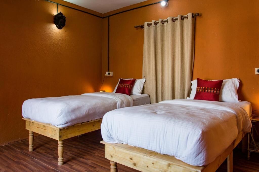 Habitación con dos camas individuales, cabeceros de madera, un ventanal con cortinas color beige y una lámpara fijada a la pared.