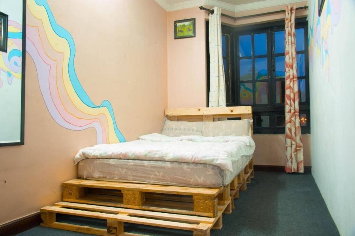A room with a bed on wooden pallets, a window with curtains, and a wall with a colorful abstract painting.