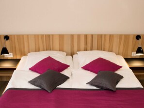 A neatly made bed with white linens, two red pillows, and two gray pillows. There are two bedside lamps on wooden nightstands. - Double room small (with balcony) - Brewery-Gasthof Pfister (Eggolsheim)
