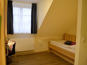 Una habitación pequeña con cama individual, techo inclinado, ventana con cortinas, un radiador y una silla con cojín estampado.