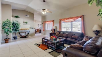 A spacious living room with a dark leather sofa, a coffee table, and a ceiling fan. There are potted plants and a dining area in the background.