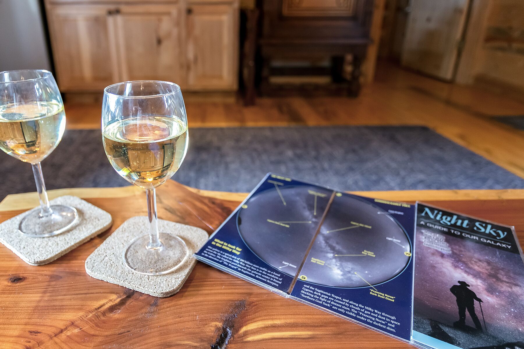 Twee glazen witte wijn op onderzetters, een sterrenkaart en een boek getiteld 'Night Sky'.