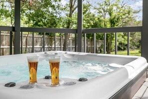 Ein Whirlpool mit zwei Biergläsern auf einem weißen Tisch, umgeben von einem Holzzaun und Bäumen.