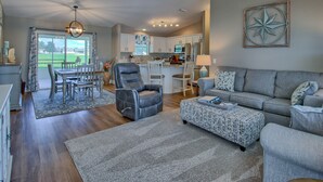 Een ruime woonkamer met een grijze bank, een leren fauteuil en een gestreept voetenbankje. Er is een plafondventilator en inbouwspots te zien.