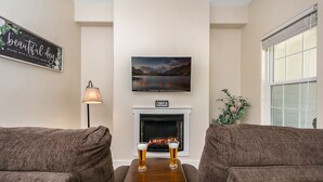 A cozy living room with a fireplace, a television mounted above it, and two armchairs facing a wooden coffee table with glasses of beer.