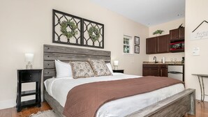A bedroom with a wooden headboard, a large bed with a brown blanket, two nightstands with lamps, a small rug, and a kitchen area with cabinets and a microwave.