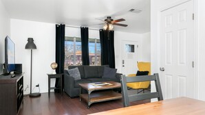 Una sala de estar moderna con un sofá gris, una mesa de centro de madera, un sillón amarillo, un televisor y un ventilador de techo.
