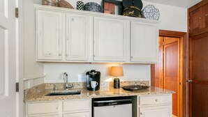 Cocina compacta con armarios blancos, lavavajillas integrado, cafetera y una lámpara sobre la encimera.