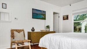A bedroom with a bed, a wooden dresser, a wicker chair, and a painting on the wall.
