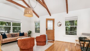 A spacious living room with a high ceiling, wooden beams, a large window, and a door with a decorative glass panel.