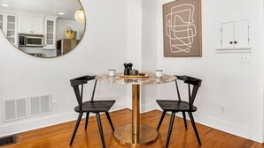 A dining area with a round table, two chairs, a hanging light, a kitchen in the background, and a framed abstract artwork on the wall.