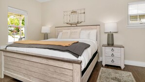 A bedroom with a wooden bed, two bedside tables with lamps, a small nightstand, and a window with blinds. - Main Street Retreat Almost Heaven Suite | King Bed | Shared Hot Tub/Pool (Fredericksburg)