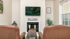 A living room with a fireplace, a television mounted above it, and two armchairs facing the fireplace. - Main Street Retreat Almost Heaven Suite | King Bed | Shared Hot Tub/Pool (Fredericksburg)