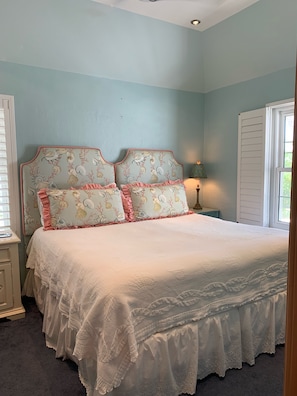 Relax in this serene bedroom with a large bed and floral-patterned headboard, set against light blue walls and a window with white shutters.