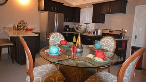 A dining area with a glass table surrounded by chairs, a kitchen in the background, and a clock on the wall. - The Monet Place (Ocho Rios)