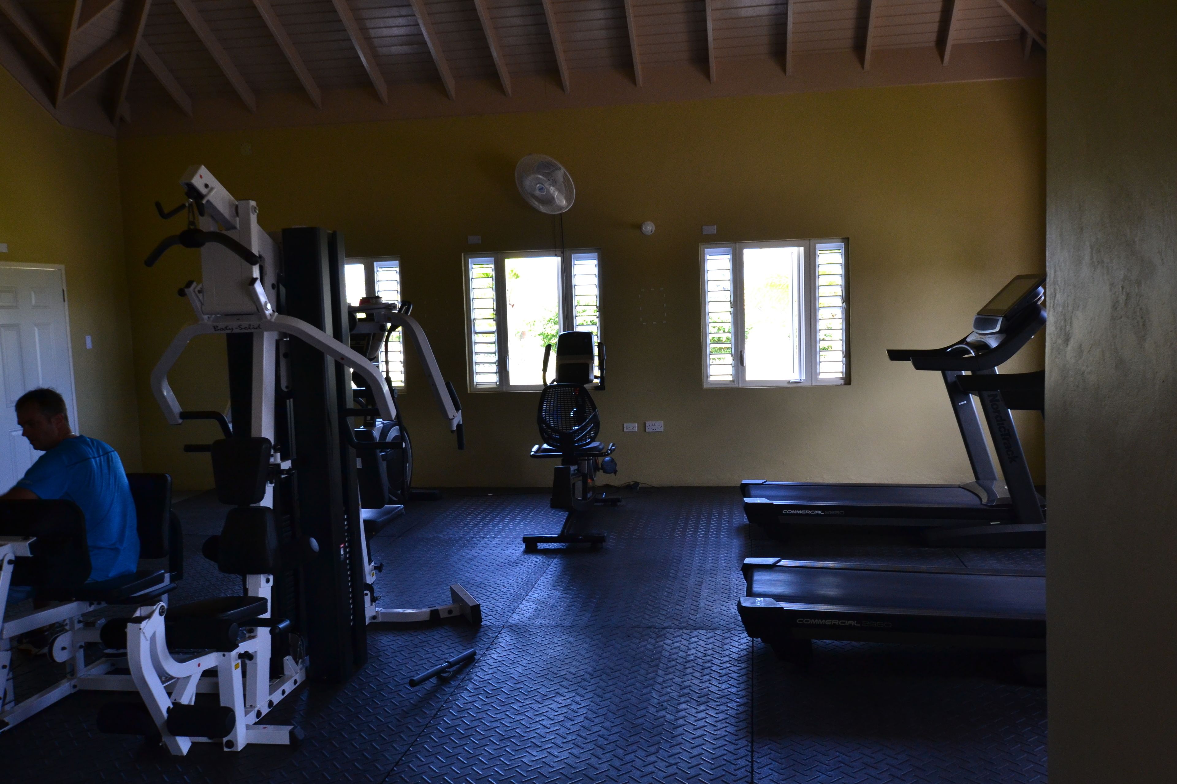 Un gimnasio con equipo de ejercicio, una cinta de correr y ventanas con persianas.