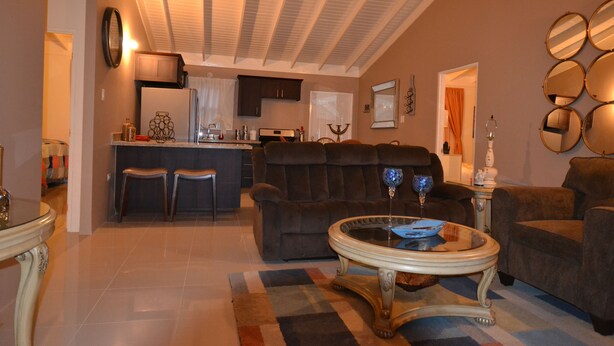 A living room with a sofa, armchairs, a coffee table, and a kitchen in the background. - The Monet Place (Ocho Rios)