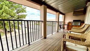 Une terrasse en bois couverte, dotée de chaises en osier et d’une table, qui offre une vue sur un parking et des arbres.