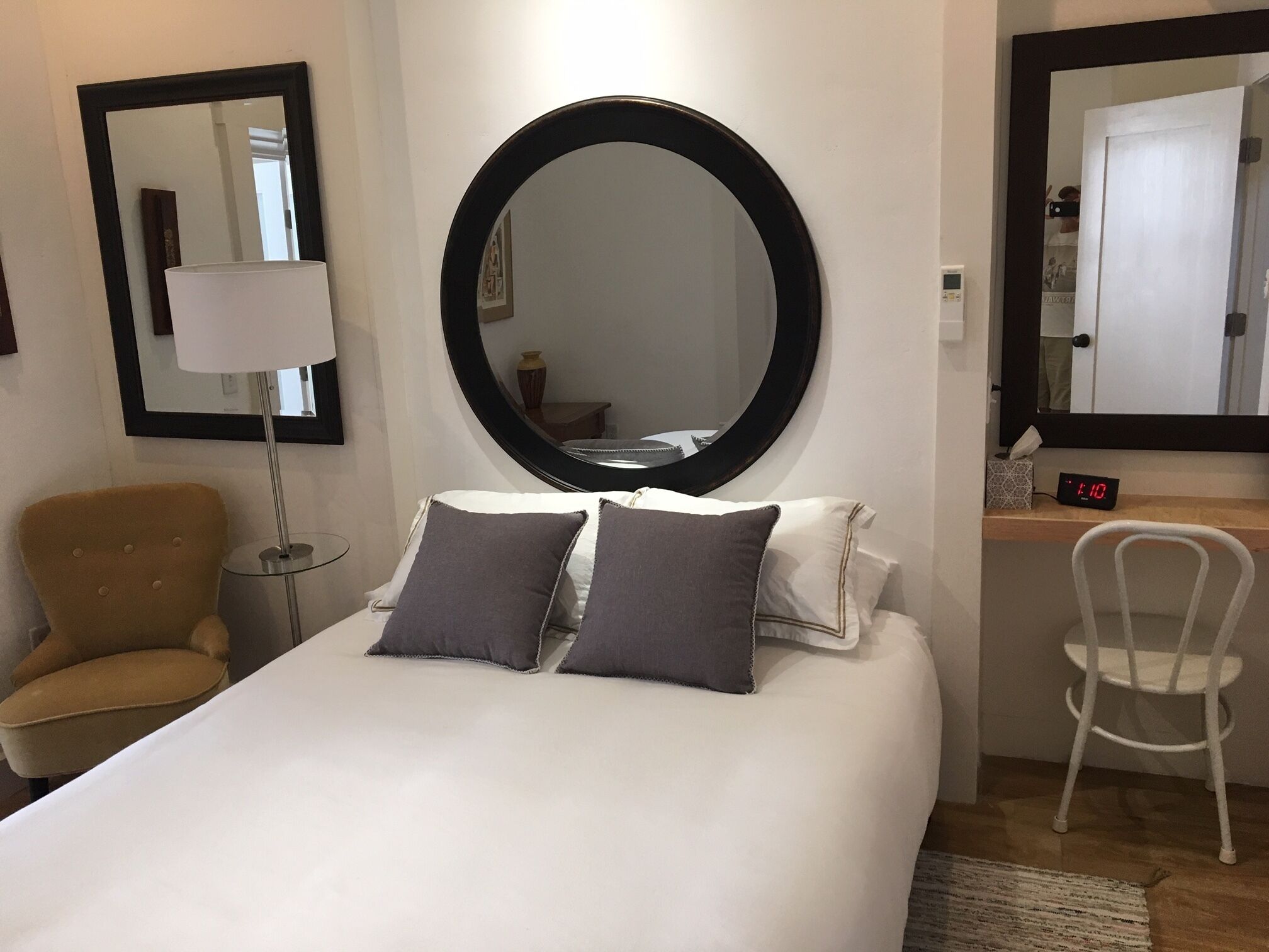 Et hotelværelse med en stor seng, to puder, et rundt spejl over sengen, en lampe, en stol og et digitalt ur.