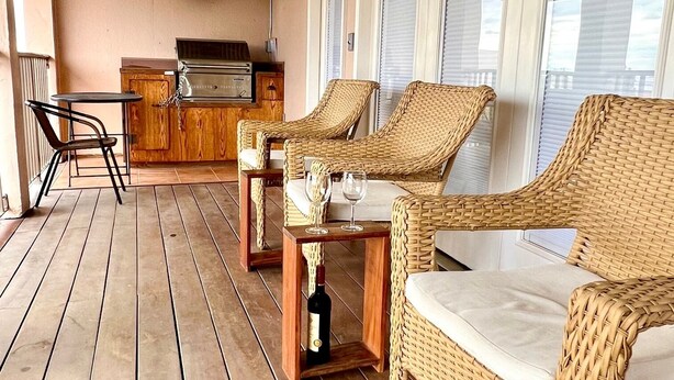 Un patio extérieur couvert, aménagé avec des meubles en osier, une table avec des verres et une bouteille, et un espace barbecue.
