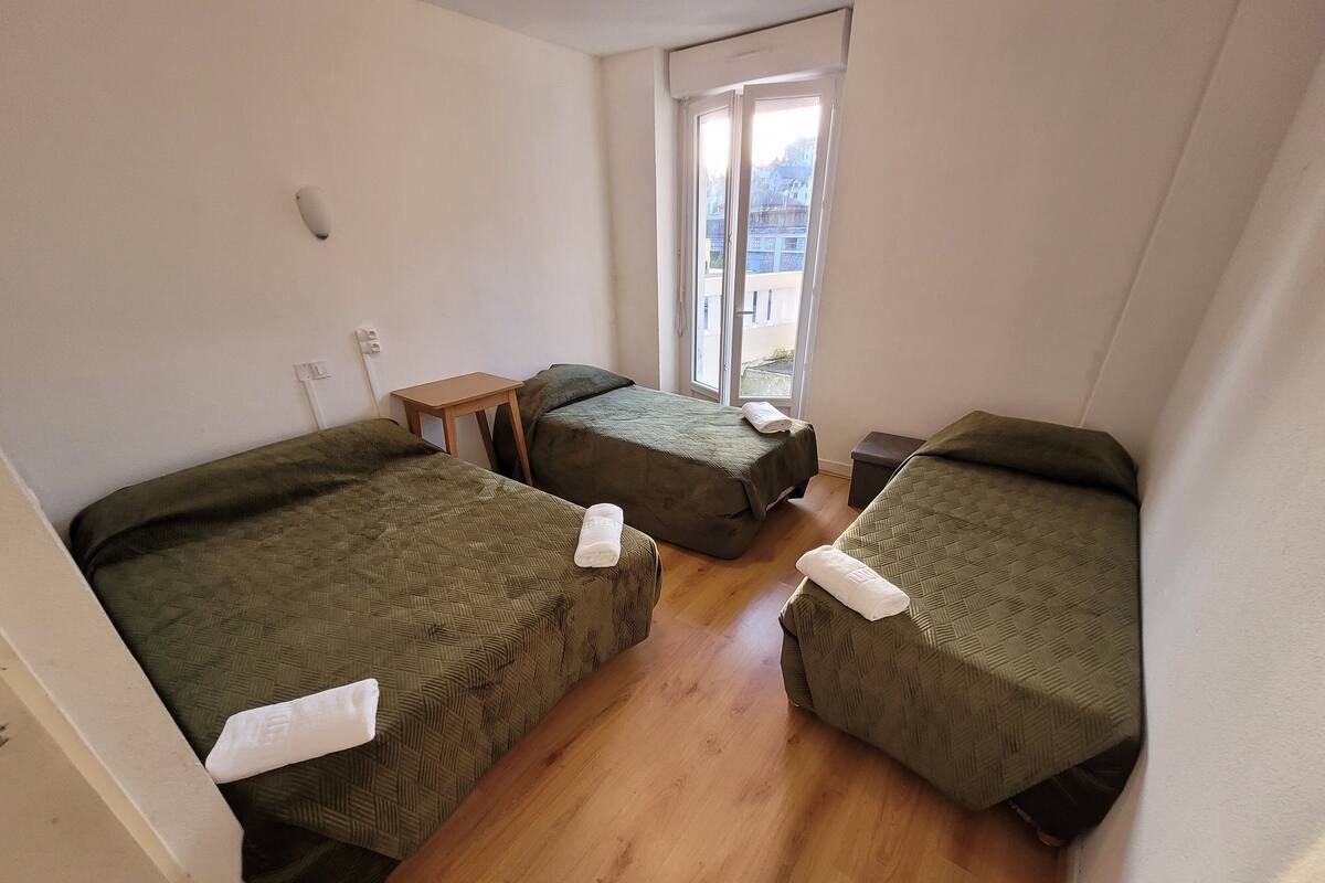 A small, clean room with two single beds, a wooden table, and a window with a view of buildings.