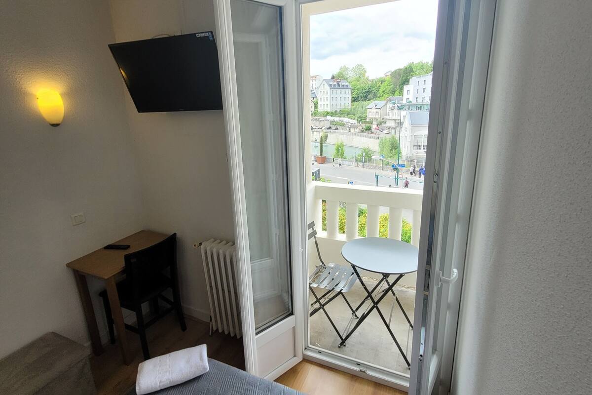A hotel room with a bed, a desk with a chair, a radiator, a TV mounted on the wall, and a balcony with a table and chairs.