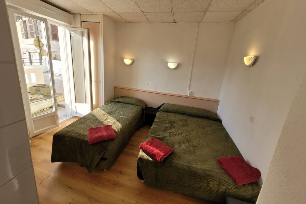 A room with two single beds, wooden flooring, a sliding glass door leading outside, and two wall-mounted light fixtures.