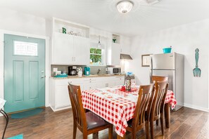 Una cocina compacta con armarios blancos, una puerta verde azulado y un comedor con mantel a cuadros.