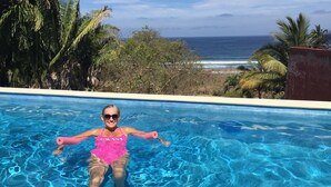 A person floating in a clear blue pool with a view of the ocean and palm trees in the background.