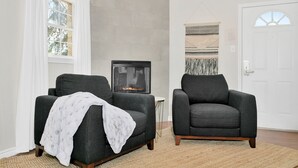 Una habitación con dos sillones grises oscuros, una mesita blanca, una chimenea, un tapiz grande tejido y una puerta blanca con un ventanal.
