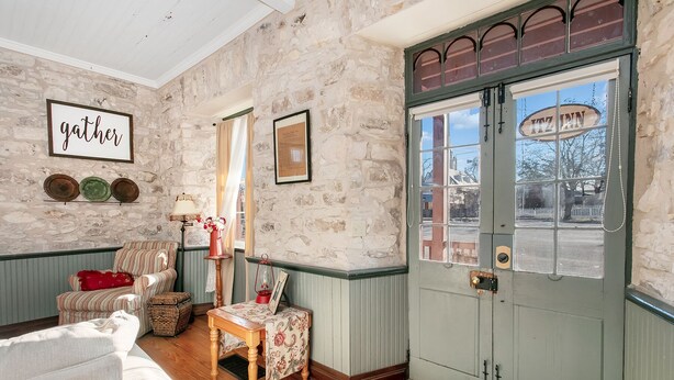 Une pièce chaleureuse avec un mur en pierre, une porte en bois ornée d’une enseigne et, au-dessus d’une étagère remplie d’assiettes, une autre enseigne indiquant « rassemblement ».
