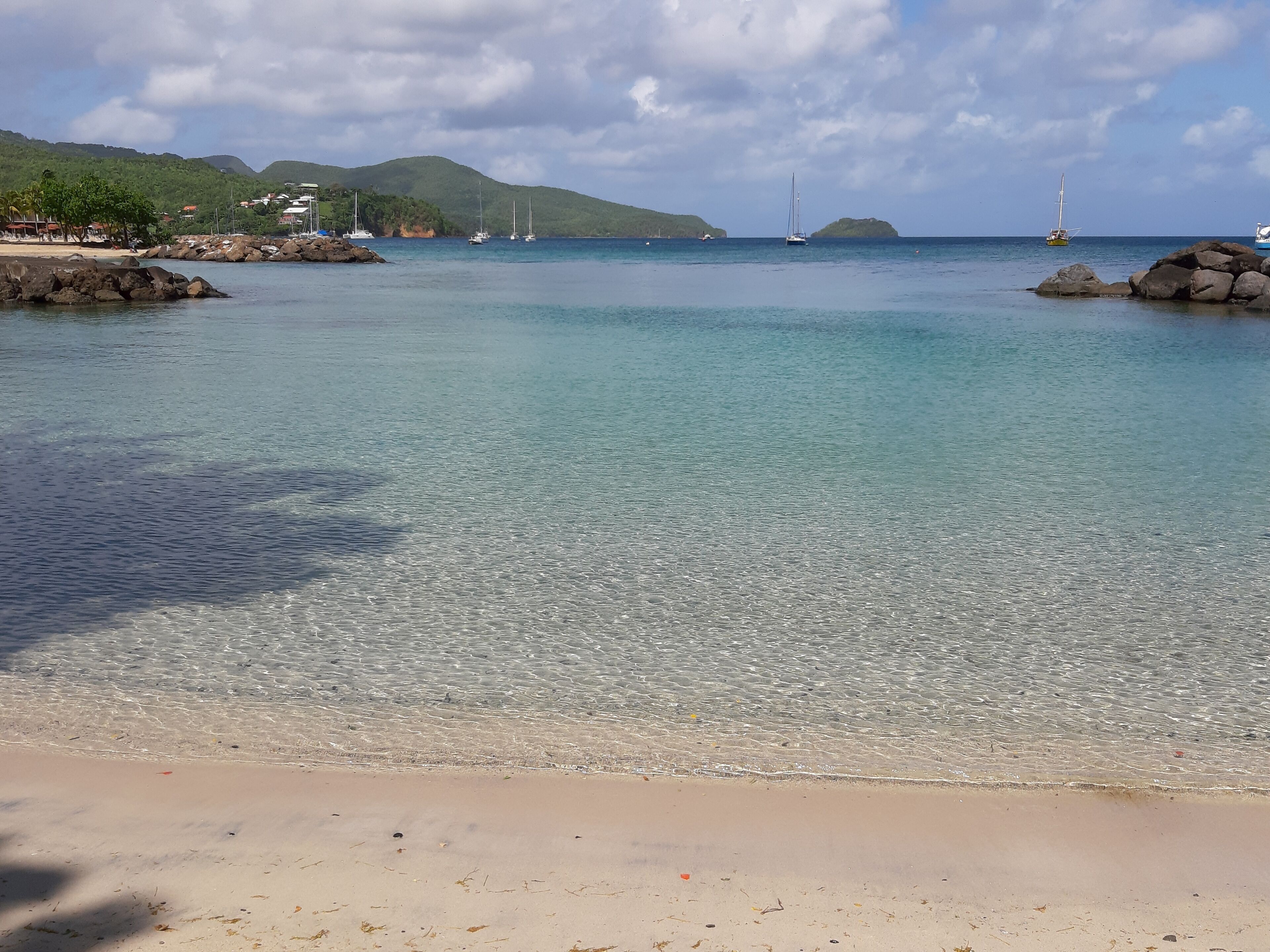 Ein Strand mit klarem Wasser, Felsen und Segelbooten in der Ferne.