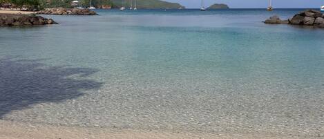 Ein Strand mit klarem Wasser, Felsen und Segelbooten in der Ferne.