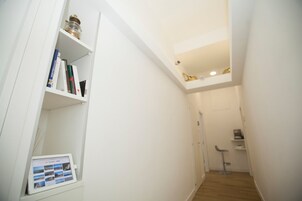 Un pasillo estrecho con paredes blancas, suelo de madera y un pequeño estante con libros y un marco digital para fotos.