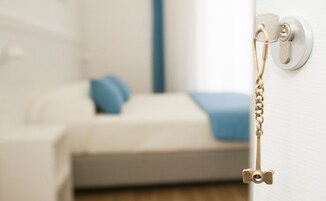 Un ojo de cerradura con una llave introducida, una puerta blanca y un fondo borroso con una cama y almohadas azules.