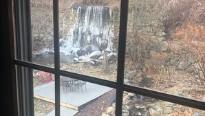 En vy över ett vattenfall genom ett fönster, med en stenmur och en träveranda med stolar och bord.