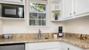 Una cocina con encimeras de granito, armarios blancos, microondas integrado, cafetera y una ventana con persianas.