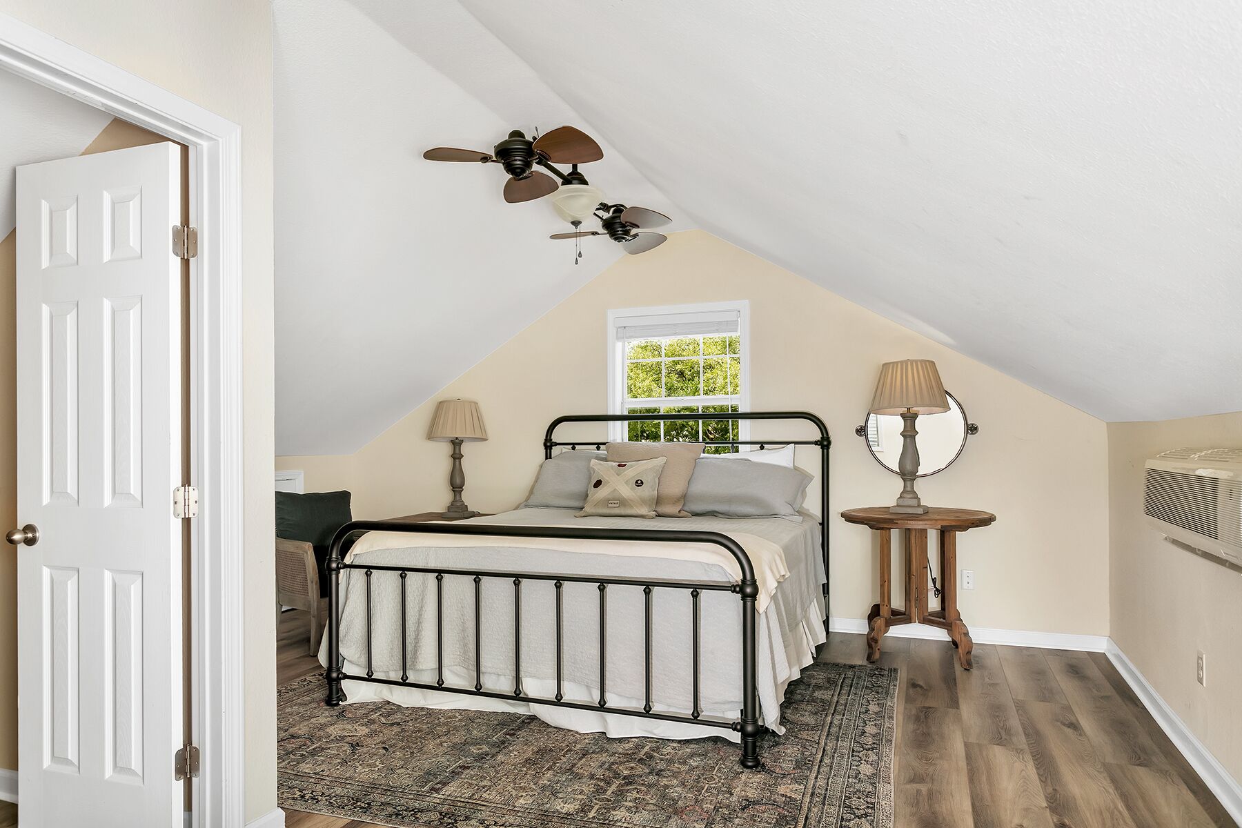 Una camera da letto con soffitto spiovente, un letto con struttura in metallo, due abat-jour, una finestra vista verde e una ventola a soffitto.