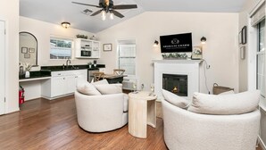 A modern living room with a fireplace, two white armchairs, a coffee table, and a kitchen area with a microwave and cabinets.