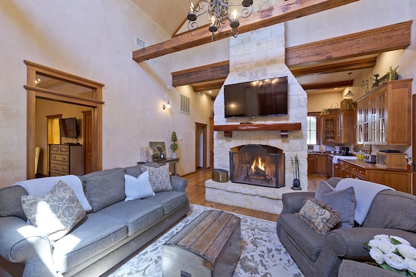A spacious living room with a fireplace, wooden beams, and a kitchen area in the background.