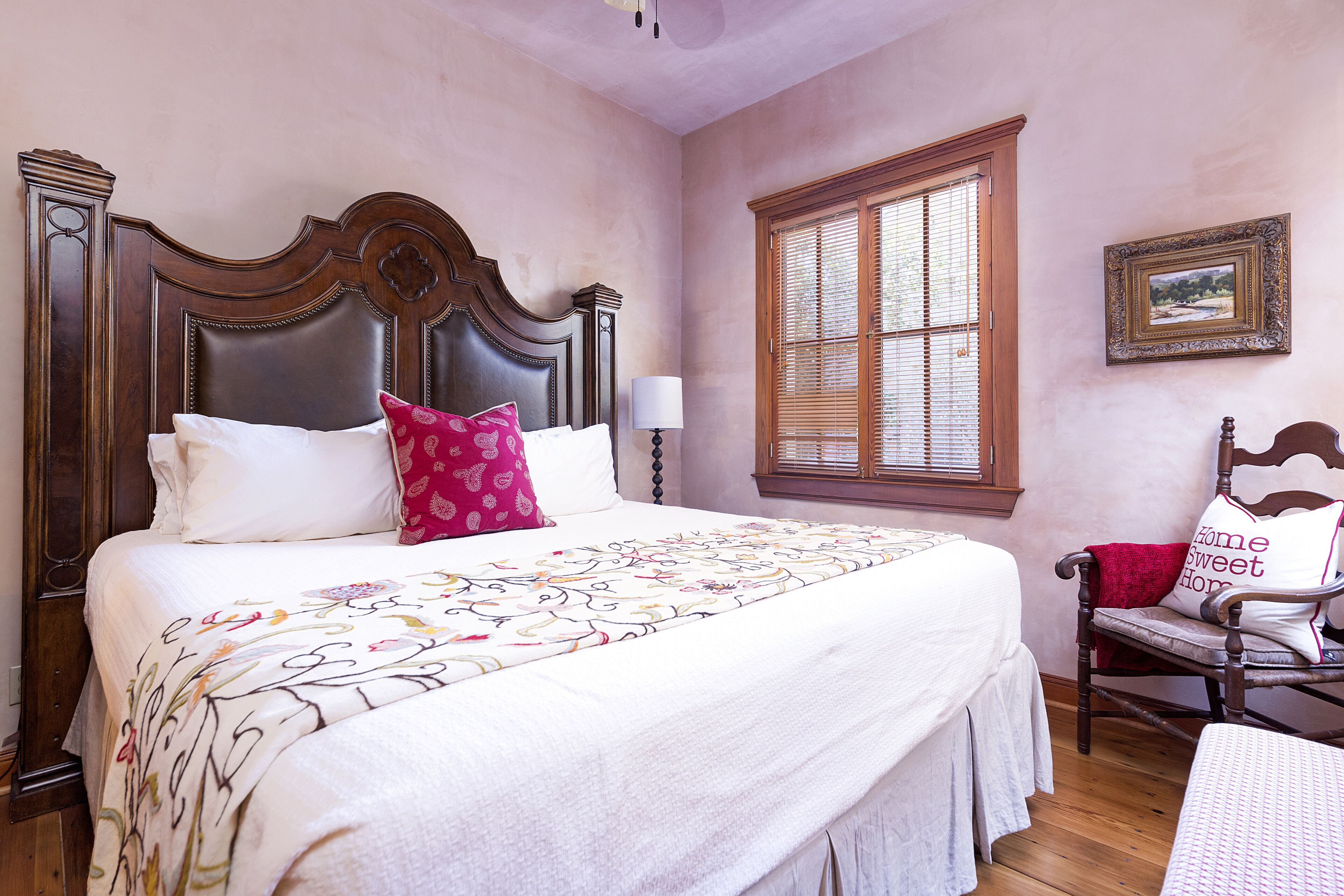 Une chambre avec un grand lit, une chaise en bois, une fenêtre avec des stores et un tableau encadré au mur.