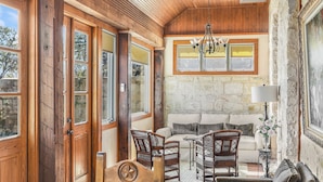 A cozy room with a stone wall, wooden ceiling, and a large window. There is a sofa, two wicker chairs, and a table with a lamp.